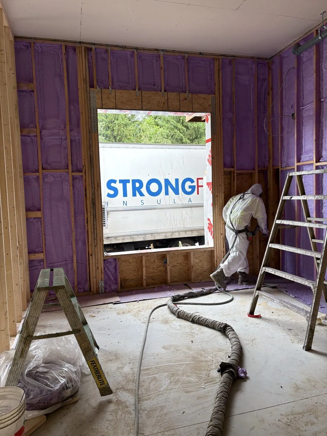 Applicator spraying foam on interior walls with the Strong Foam truck visible through the window